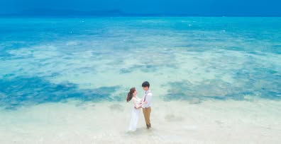 石垣島の「電信屋ビーチ」にて、透明度抜群のクリスタルブルーの海に足元を浸しながら見つめ合う新郎新婦。波打ち際の透き通るような白砂と、幾重にも重なる青のグラデーションが美しい海が広がる、幻想的で清涼感あふれるウエディングフォト。