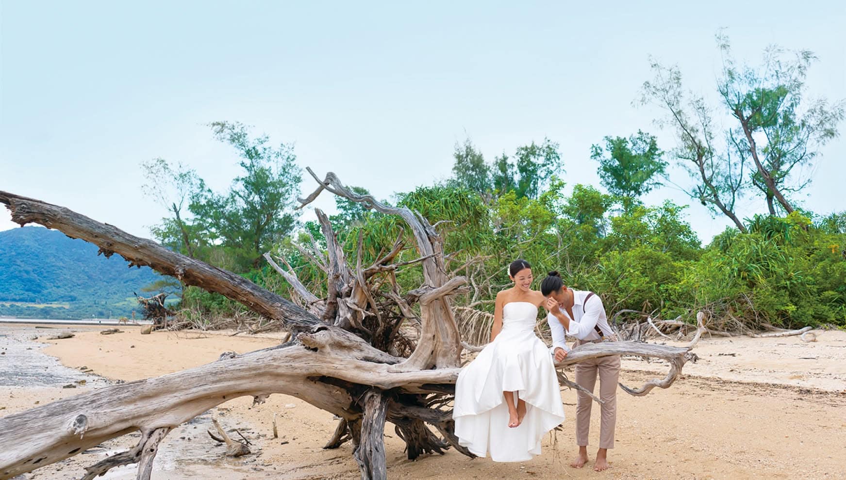 石垣島の「名蔵大橋下ビーチ」に横たわる巨大な流木に座り、楽しげに笑い合う新郎新婦。背景には名蔵湾の穏やかな海と山並みが広がり、自然が造り出したダイナミックなオブジェと、裸足でリラックスしたスタイルの二人が調和する、開放感あふれるウエディングフォト。