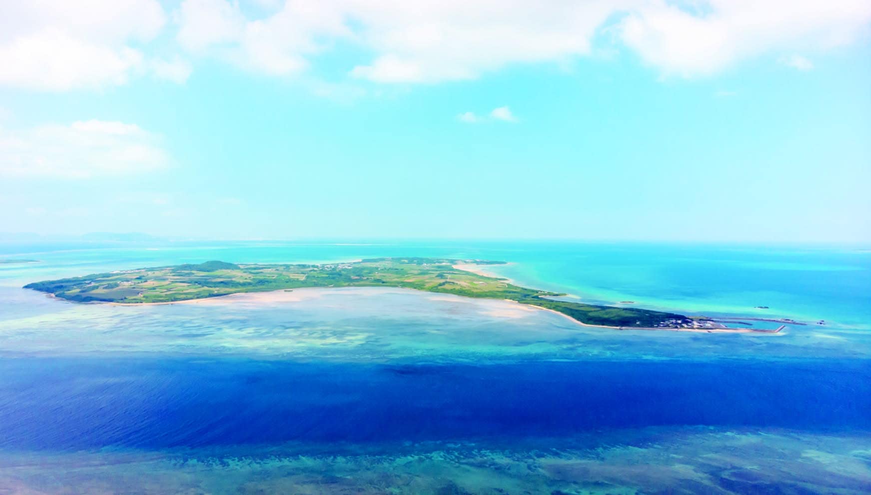 八重山諸島に位置する「小浜島（こはまじま）」の壮大な空撮。コバルトブルーからエメラルドグリーンへとグラデーションを描く透明度の高い海と、島を囲む広大なサンゴ礁のリーフ。沖縄リゾートの美しさを象徴する、フォトウエディングの舞台としての魅力を伝える一枚。