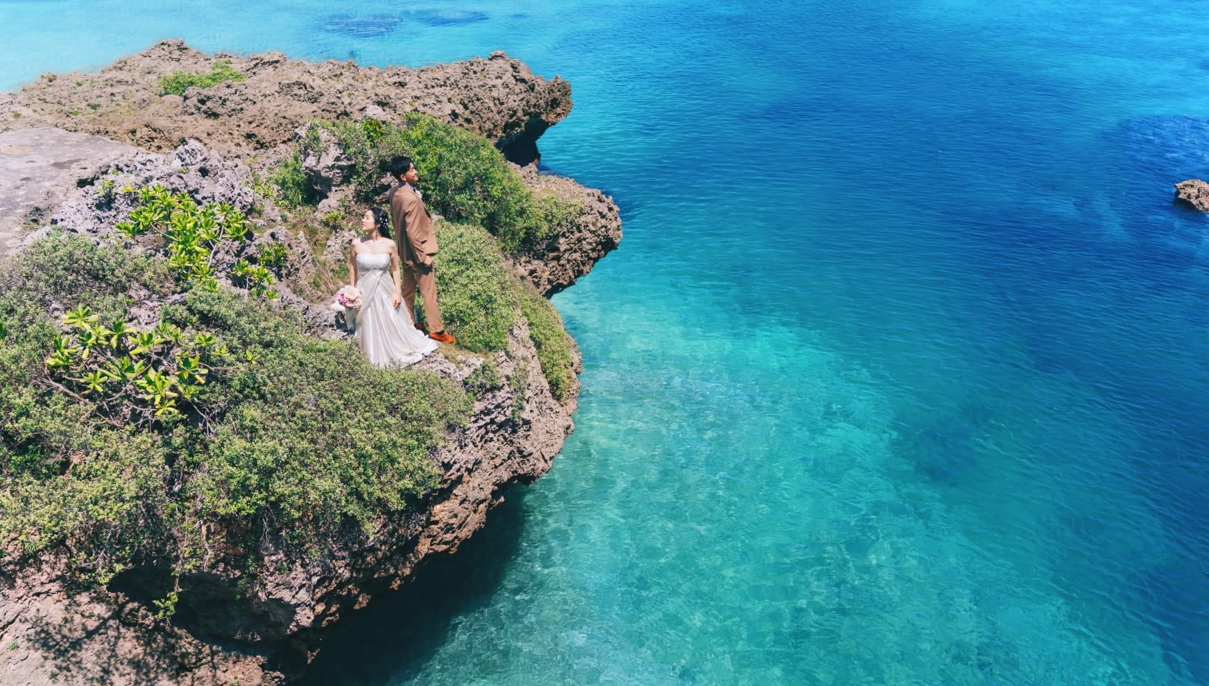 宮古島の「イムギャーマリンガーデン」にて、断崖絶壁のような岩場から息をのむほど美しいコバルトブルーの海を見下ろす新郎新婦。ダイナミックな海岸線と透明度の高い海が広がる絶景を背景に、ブラウンのスーツを纏った新郎とウエディングドレス姿の新婦が佇む、壮大なロケーションフォト。