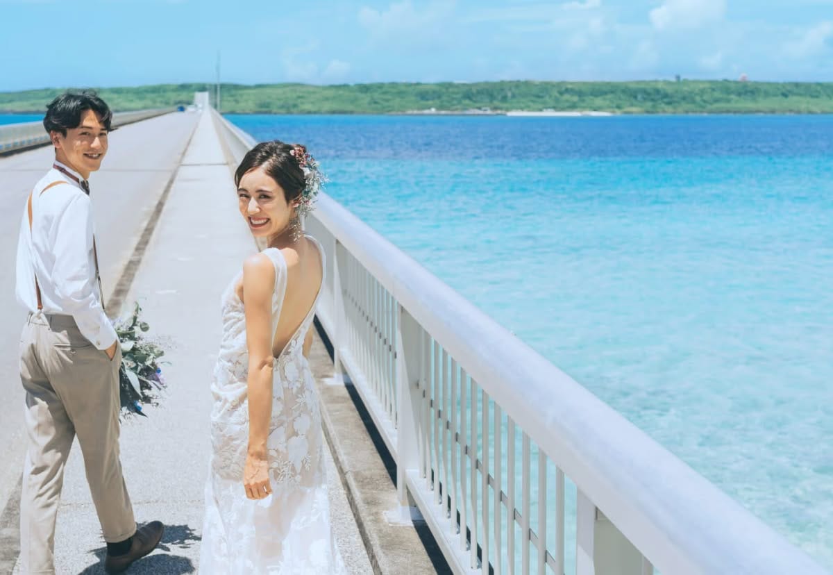 宮古島と来間島を結ぶ「来間大橋」の上で、エメラルドグリーンの美しい海をバックに笑顔で振り返る新郎新婦。青い海と空がどこまでも続く絶景の中、サスペンダー姿の新郎と、背中が大きく開いたレースのドレスを纏った新婦が、開放感あふれるロケーションを楽しむ爽やかなウエディングフォト。