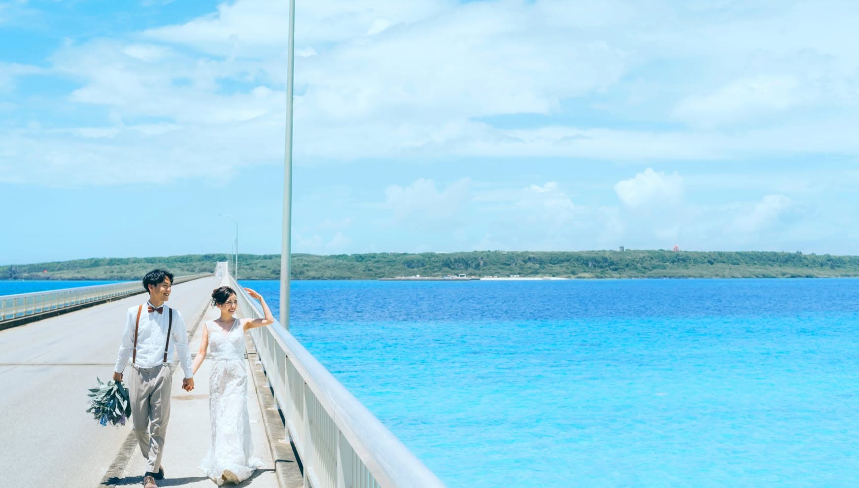 宮古島と来間島を結ぶ来間大橋の上で、真っ青な海をバックに手をつないで歩く新郎新婦。サスペンダー姿の新郎と、総レースのウエディングドレスを纏った新婦が、心地よい海風を感じながら笑顔を見せる、開放感に満ちたロケーションフォト。