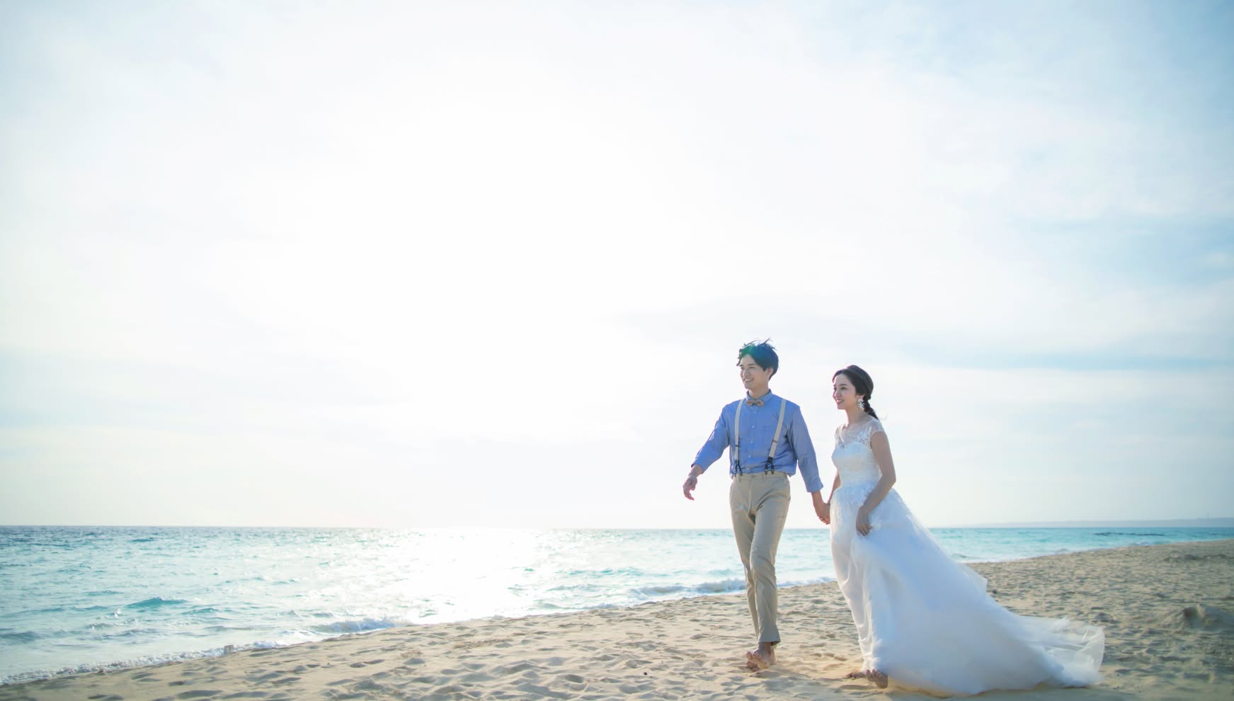 宮古島の与那覇前浜ビーチにて、東洋一とも称される白い砂浜と透き通る海をバックに、手を繋いで歩く新郎新婦。柔らかな逆光が二人を包み込み、ブルーのシャツにサスペンダー姿の新郎と、風に美しくなびくウエディングドレスを纏った新婦が、幸せそうに微笑むロマンチックなカット。