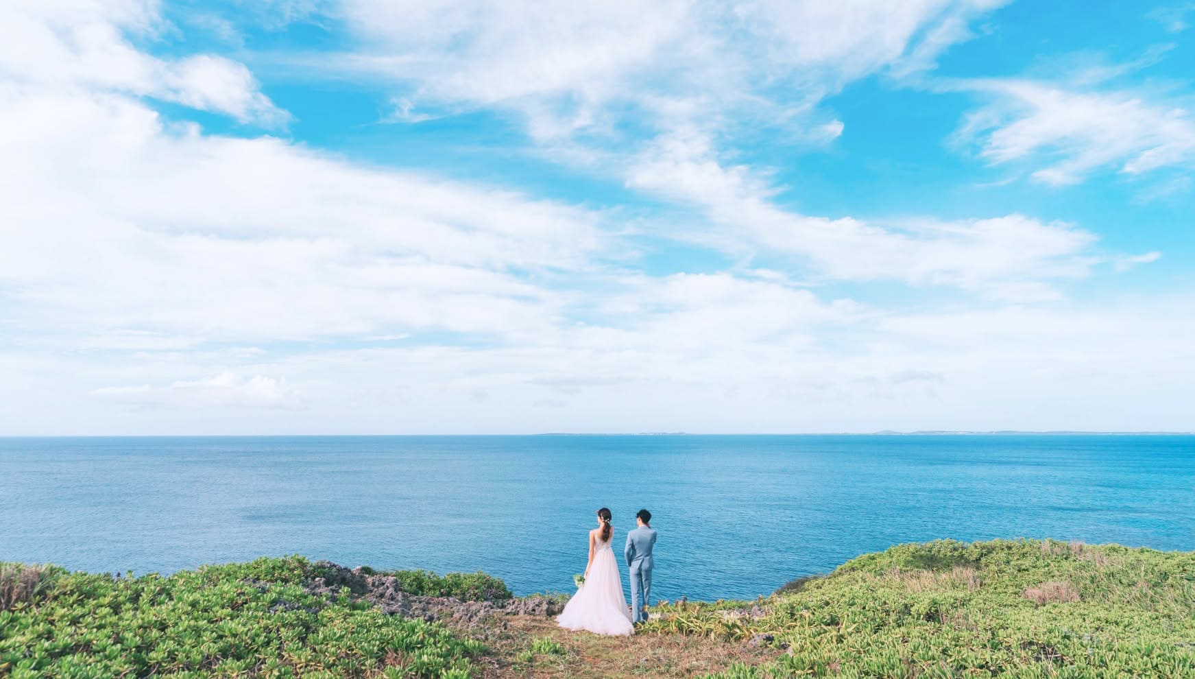 宮古島の絶景スポット「ムイガー断崖」の切り立った崖の上から、果てしなく続く紺碧の海を眺める新郎新婦のバックスタイル。鮮やかな緑の植生と、地平線まで広がる青い空と海が溶け合うような、壮大なスケールのロケーションフォト。