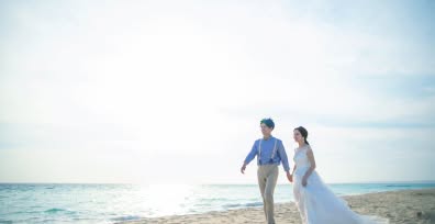 きらめく青い海を背景に、白い砂浜を仲良く歩く新郎新婦のウエディングフォト。新婦は軽やかな白いドレス、新郎は青いシャツにサスペンダーを合わせたカジュアルな装いで、明るい太陽の下、開放感あふれるビーチロケーションを楽しむ二人を捉えた一枚。