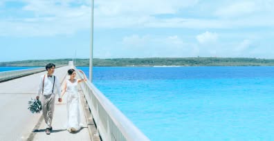 果てしなく続くエメラルドグリーンの海を背景に、真っ直ぐな橋の上を仲良く手をつないで歩く新郎新婦のウエディングフォト。白いレースのドレスを着た新婦と、サスペンダー姿のカジュアルな新郎が、爽やかな海風を感じながら笑顔を見せる開放的な一枚です。