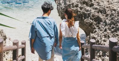 青いシャツとワンピースの私服スタイルに着替えた新郎新婦が、手すりのある階段を降りて、岩場に囲まれた隠れ家のような美しいビーチへと向かう、旅のワクワク感あふれる後ろ姿。