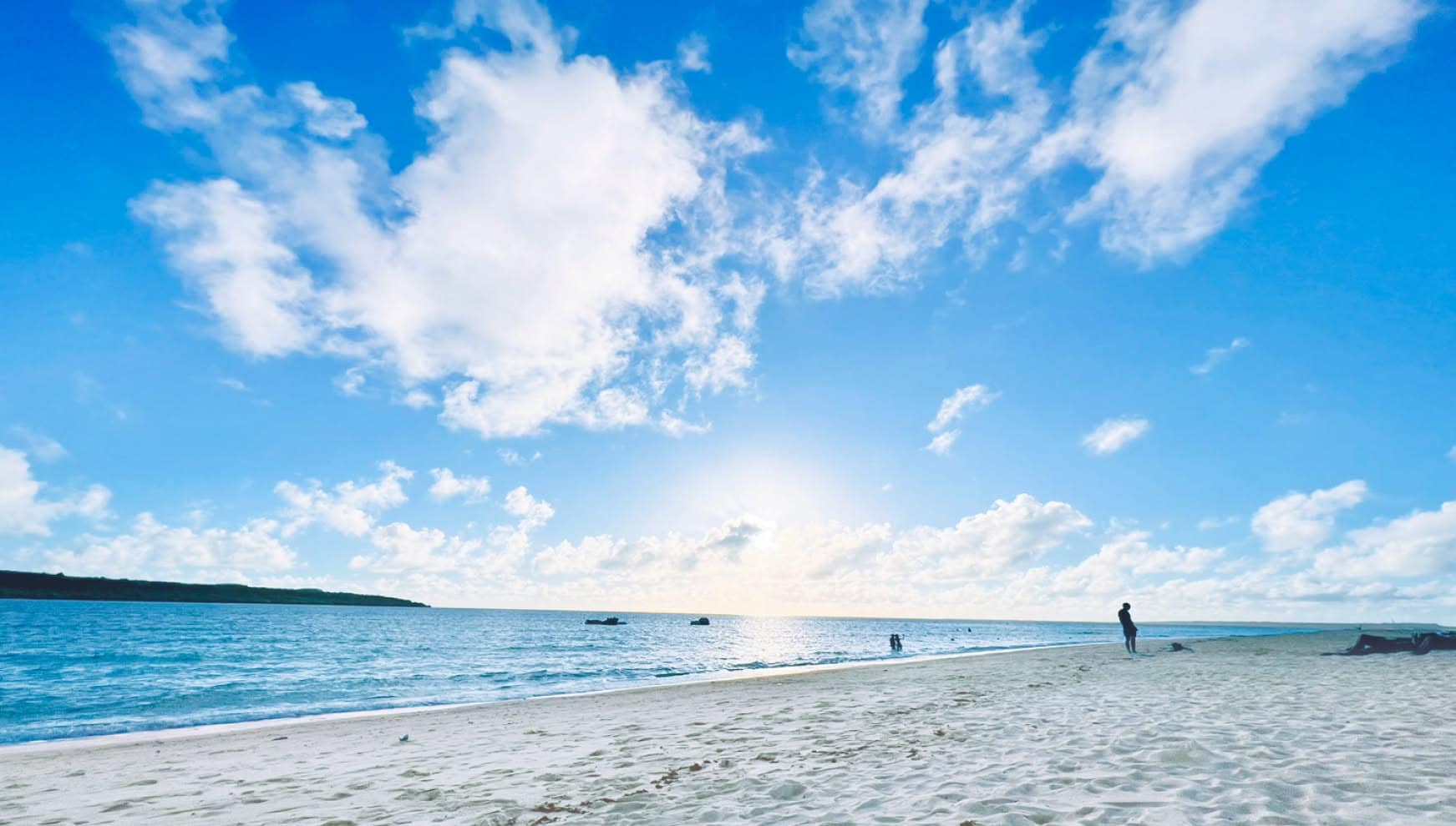 宮古島の市街地近くにあるパイナガマビーチ周辺の、白く美しい砂浜と穏やかな海。澄み渡る青い空に広がる白い雲と、水平線から差し込む柔らかな光が、リゾート地ならではの開放的な雰囲気を醸し出す絶景のロケーション。