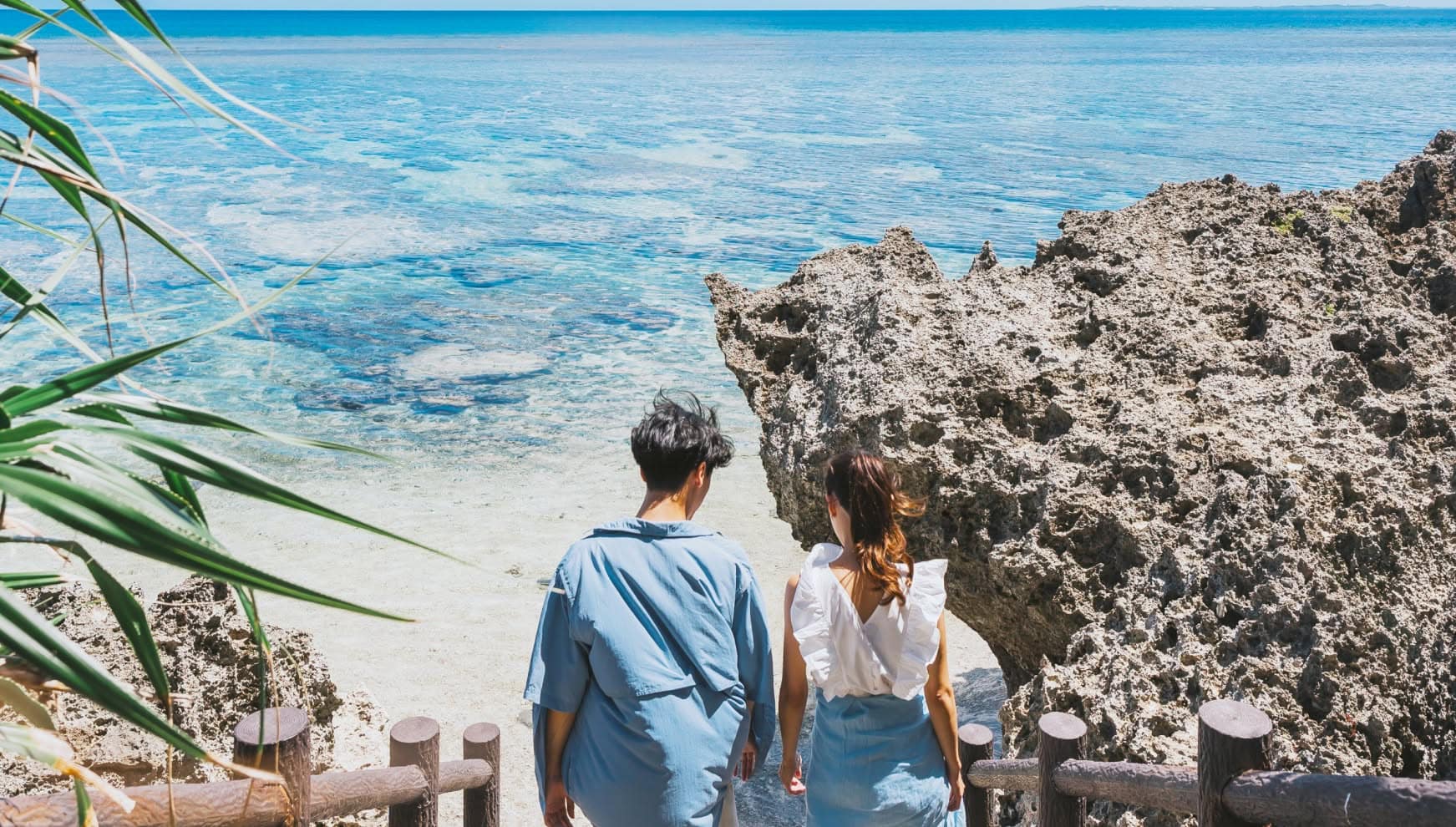 宮古島（池間島）の隠れ家的スポット「シンビジ」にて、岩場の階段からエメラルドグリーンの美しい海を見下ろす新郎新婦のバックスタイル。透明度の高い海と複雑な岩礁が織りなす絶景を背に、カジュアルな装いの二人が寄り添う、旅の情緒あふれるウエディングフォト。