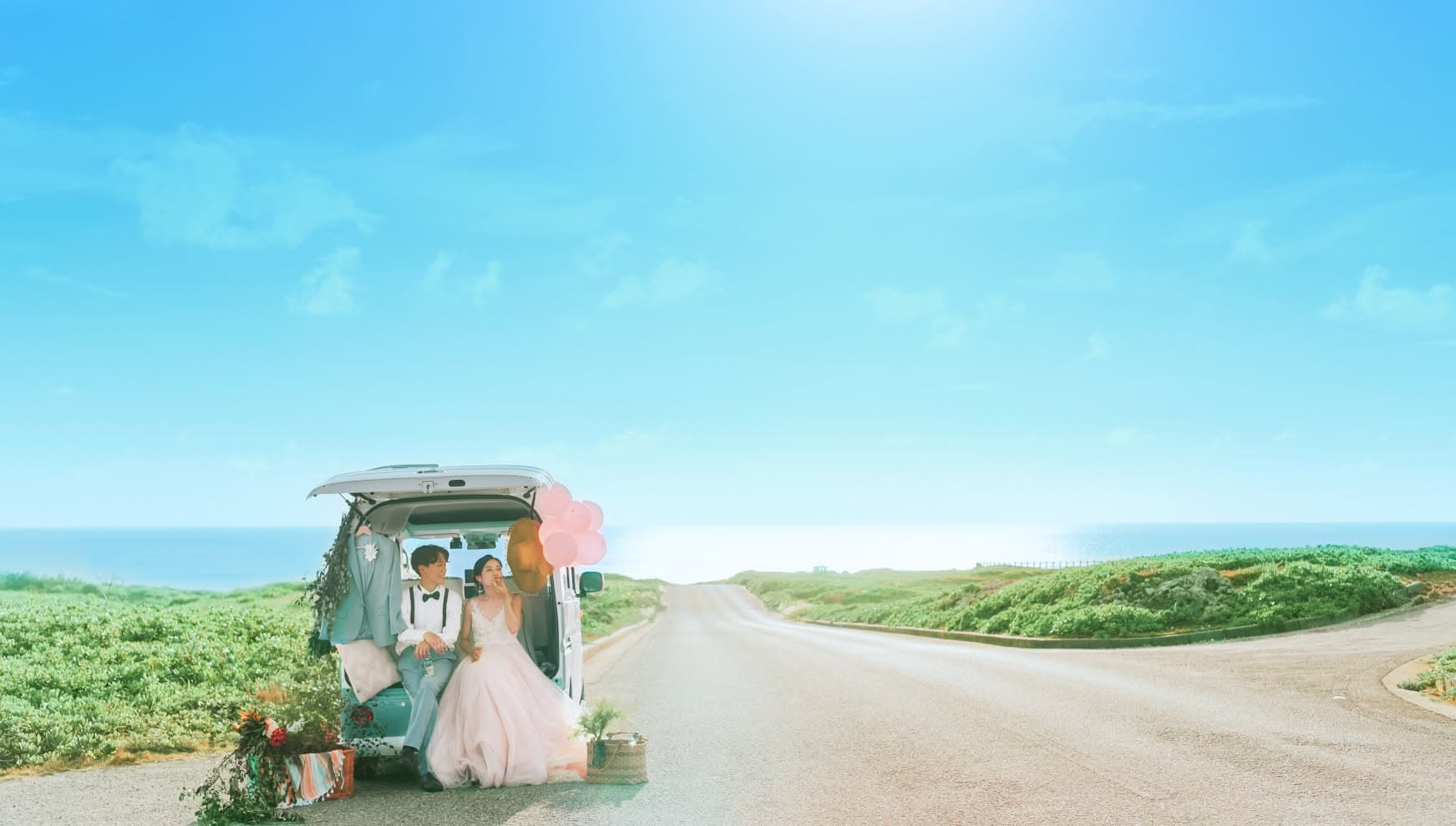 宮古島（伊良部島）の白鳥崎 西海岸公園にて、海へと続く一本道を背景に、車のバックドアを開けて座る新郎新婦。ピンクの風船や麦わら帽子、バスケットを飾り、広大な空と緑に囲まれた開放的なロケーションで楽しむ、おしゃれなバンのウエディングフォト。
