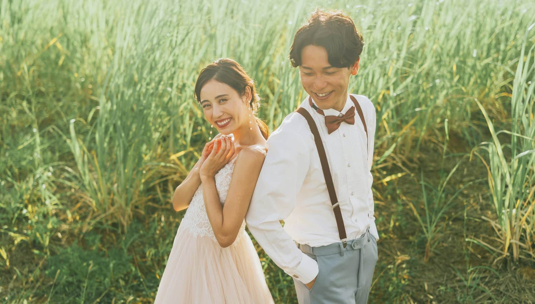 宮古島の「ウージ（サトウキビ）」畑を背景に、背中合わせで笑顔を見せる新郎新婦。柔らかな夕日の光に包まれた緑豊かなロケーションで、サスペンダー姿の新郎とナチュラルなウエディングドレス姿の新婦が、沖縄らしいのどかな風景に溶け込む温かみのある一枚。