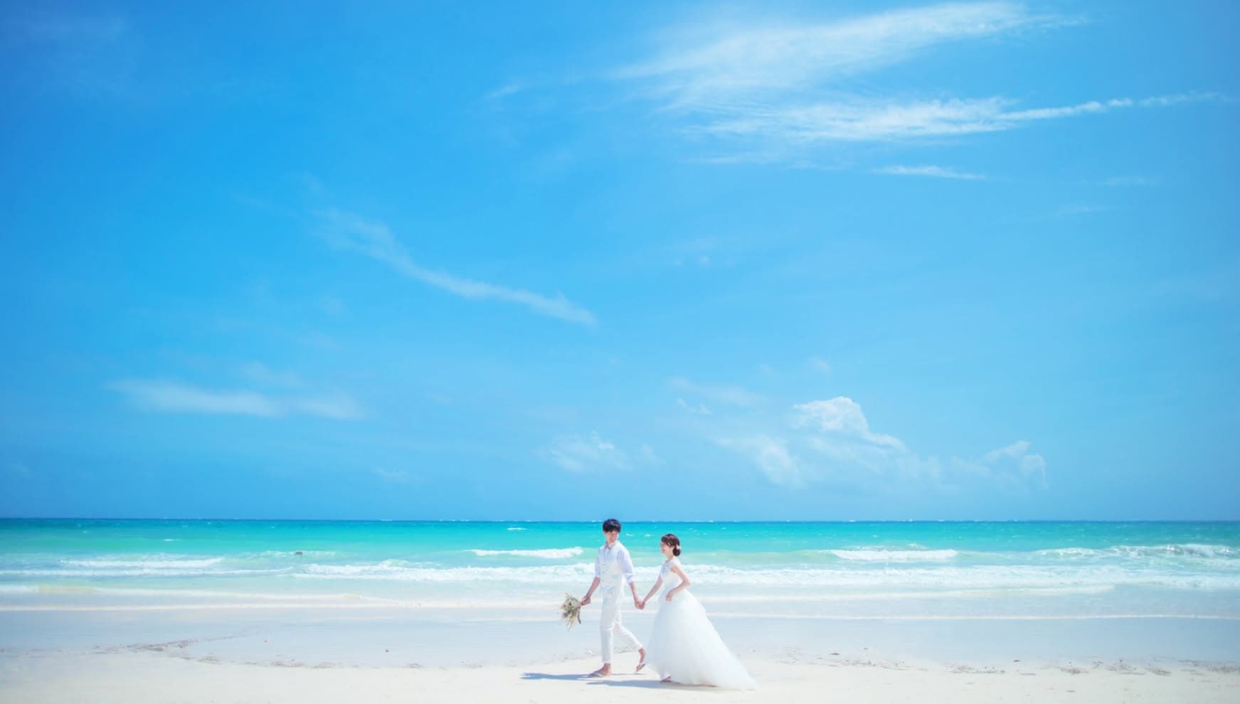 宮古島（伊良部島）の渡口の浜にて、パウダースノーのような真っ白な砂浜と透き通る宮古ブルーの海をバックに、手をつないで歩く新郎新婦。どこまでも続く青い空と白い波打ち際が美しい、開放感あふれるビーチ撮影でのウエディングフォト。