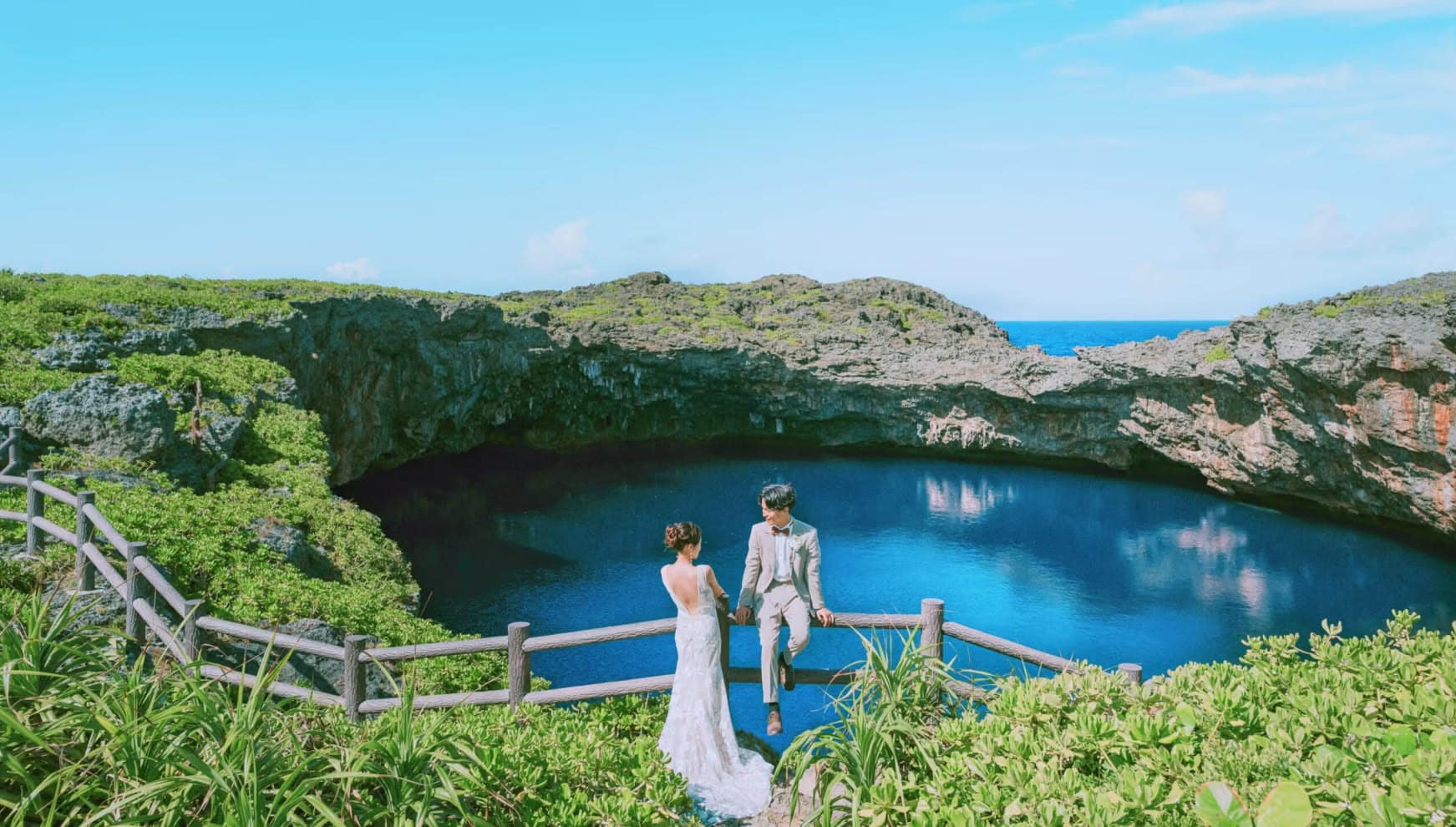 宮古島（下地島）のパワースポット「通り池」の深い紺色の水面を背景に、木製の柵に腰掛ける新郎と隣に立つ新婦。周囲を囲む荒々しい岩肌と豊かな緑、そしてどこまでも続く青い空が、神秘的でダイナミックなロケーションを演出するウエディングフォト。