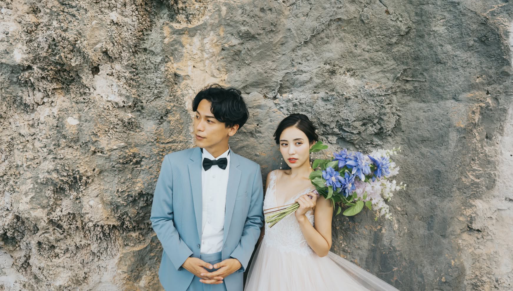 宮古島（伊良部島）の巨大な奇岩「ヤマトブー大岩」の迫力ある岩肌を背に、クールな表情で佇む新郎新婦。ライトブルーのタキシードを着た新郎と、青い花をアクセントにしたボリュームのあるブーケを持つウエディングドレス姿の新婦が、自然の造形美の中で際立つドラマチックな一枚。