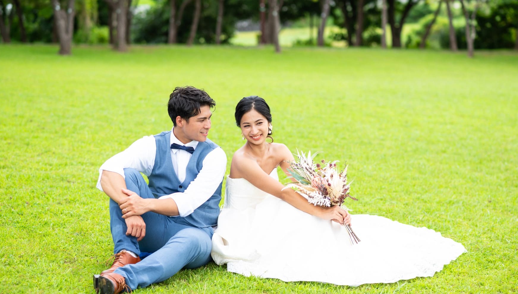 沖縄本島の21世紀の森公園にて、鮮やかな緑の芝生に座り寄り添う新郎新婦。青いベストを着用したカジュアルなスタイルの新郎と、ドライフラワーのブーケを手に持ち笑顔を浮かべるウエディングドレス姿の新婦の、穏やかな空気感が伝わるロケーションフォト。
