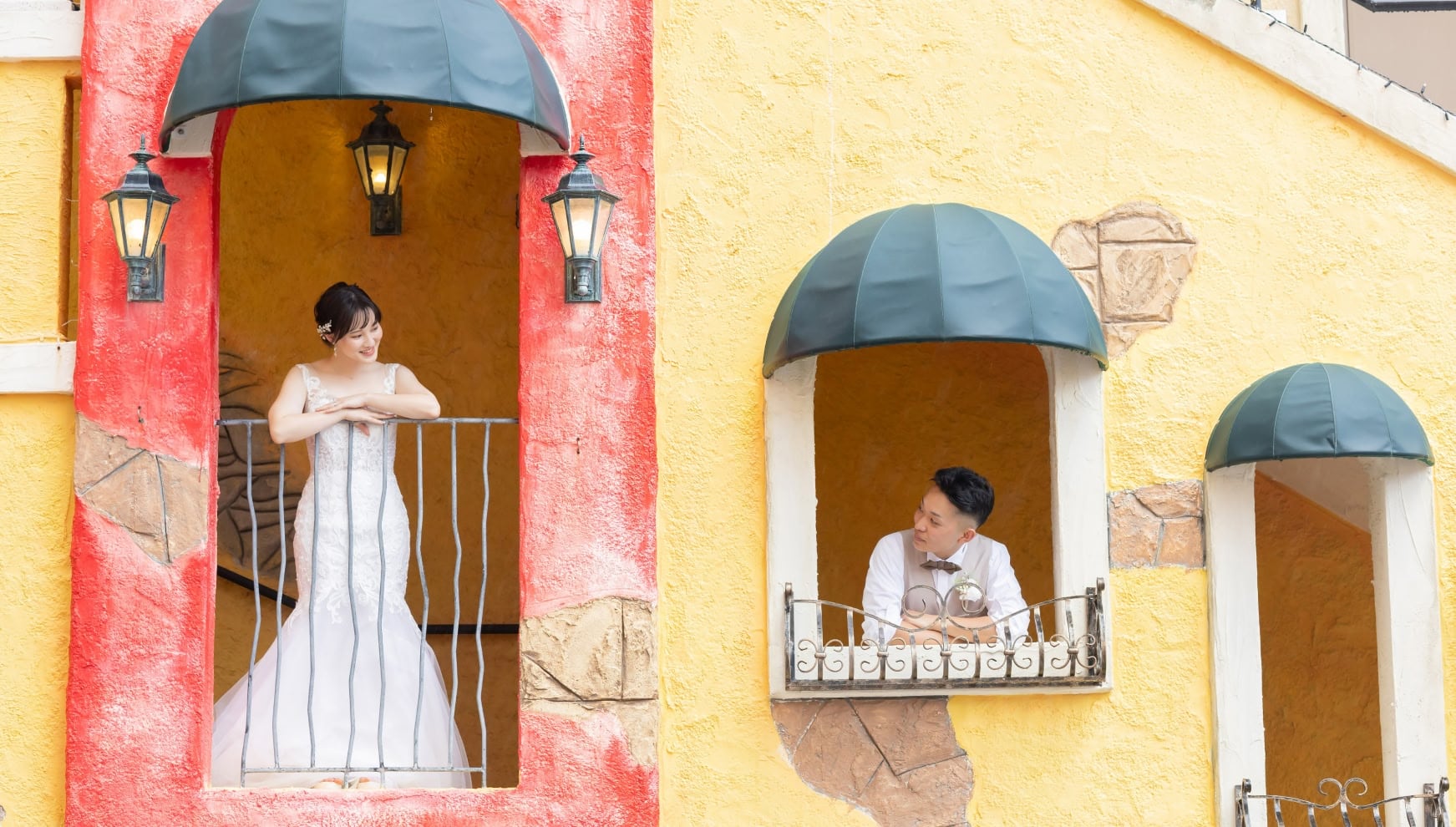 沖縄本島の北谷町にある美浜アメリカンビレッジにて、カラフルな洋風の建物の窓辺に立つ新郎新婦。マーメイドラインのウエディングドレスを着た花嫁と、隣の窓から見つめる新郎が、まるで映画のワンシーンのような世界観を演出するウエディングフォト。