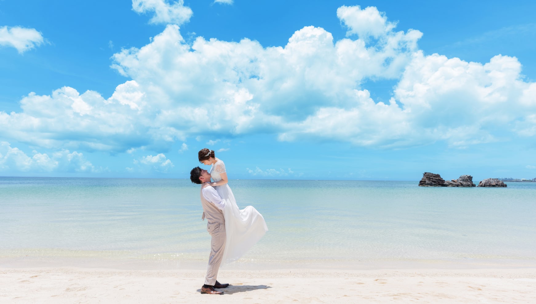 沖縄本島のアラハビーチにて、青い海と広大な空を背景に、新郎が新婦を軽やかに抱きかかえるウエディングフォト。白い砂浜と澄み渡る水平線、立体感のある白い雲が、リゾート感あふれるドラマチックなロケーション撮影を演出している。