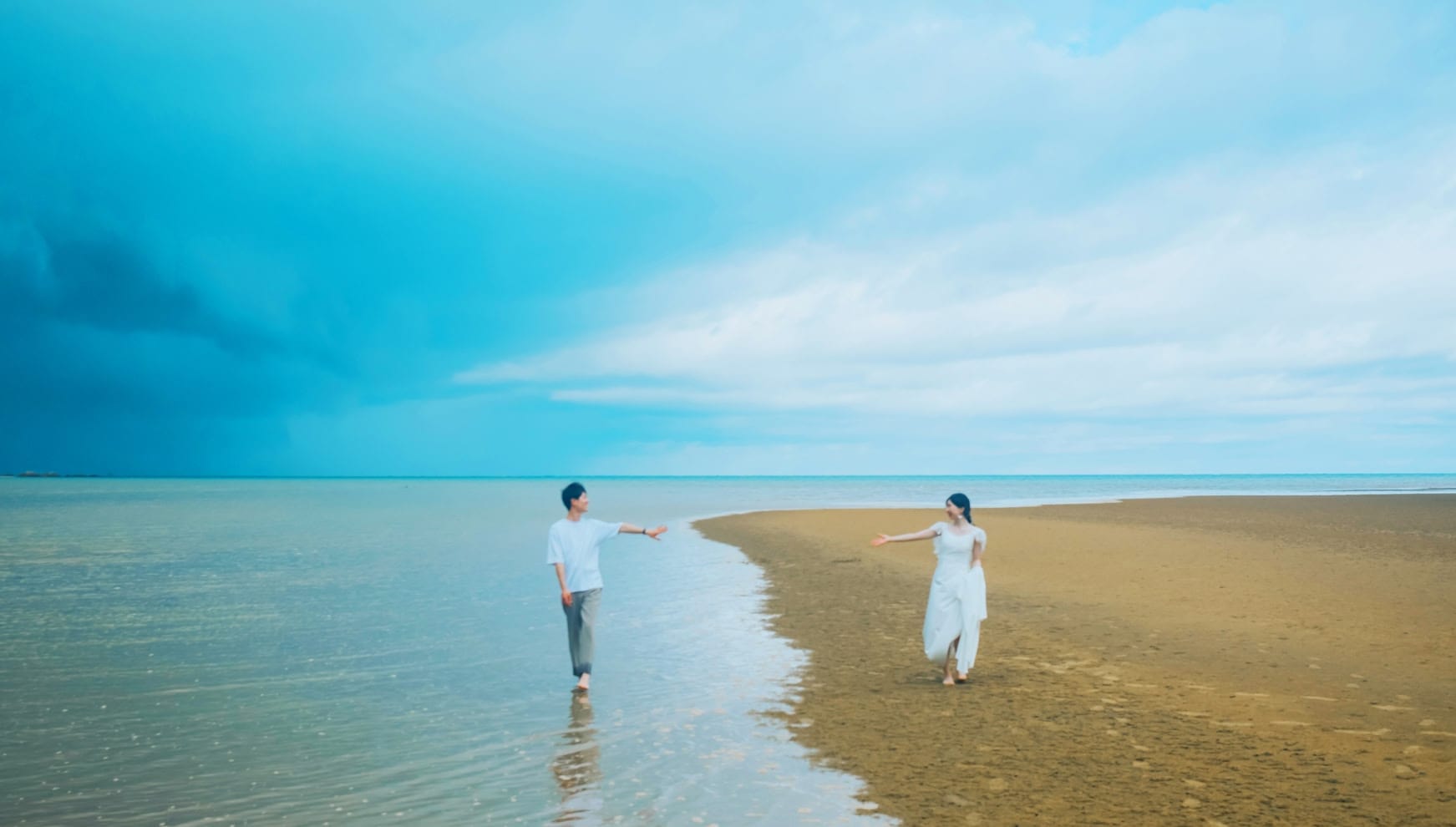 沖縄本島の潟原の干潟にて、潮が引いた広大な砂州を歩く新郎新婦。青い空と海が溶け合うような幻想的なロケーションの中、白いワンピースドレスの花嫁とカジュアルなシャツスタイルの新郎が、手を取り合おうと歩み寄る自然体なウエディングフォト。