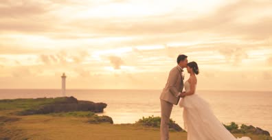 夕暮れ時の美しい海岸沿いで、新郎が新婦の額に優しくキスをするロマンチックなウエディングフォト。遠くに白い灯台が見える広大な岬を舞台に、黄金色の空と海が二人を包み込む、幻想的なサンセットロケーションでの一枚です。