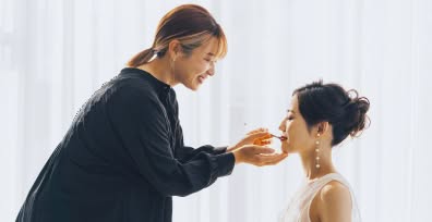 プロのヘアメイクアーティストが新婦にリップを施しているメイクシーン。清潔感のある明るい室内で、これから始まる撮影に向けて期待が高まる準備風景。