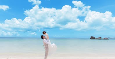 沖縄本島の真っ青な空と穏やかなビーチを背景に、新郎が新婦を軽々と抱きかかえるダイナミックな抱っこショット。白い砂浜と透き通るエメラルドグリーンの海、そして鮮やかな入道雲が広がる絶景の中での、開放感あふれるフォトウエディングの撮影風景。