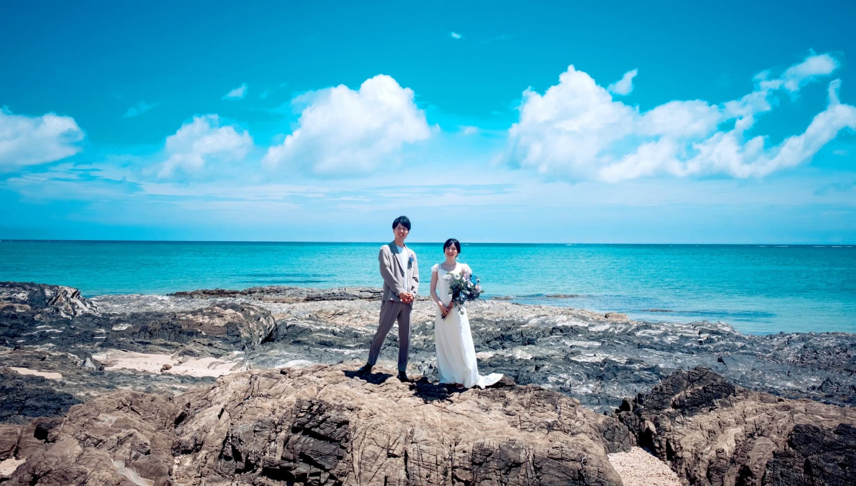 沖縄本島のシーグラスビーチにあるゴツゴツとした岩場に立ち、エメラルドグリーンの海を背景に並ぶ新郎新婦。澄み渡る青い空と白い雲の下、落ち着いたグレーのスーツを着た新郎と、ボリュームのあるブーケを手にしたウエディングドレス姿の新婦の全身カット。
