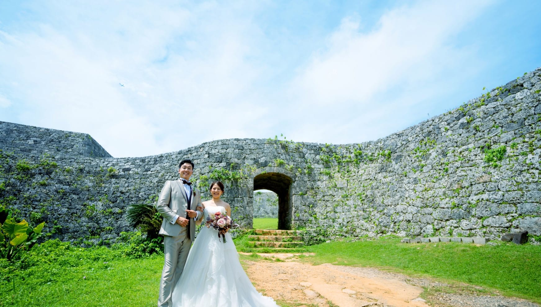 世界遺産の座喜味城跡にて、歴史を感じさせる美しい曲線を描く石垣を背景に笑顔で並ぶ新郎新婦。青い空と緑の芝生に、光沢のあるシャンパンゴールドのタキシードと純白のウエディングドレスが映える、沖縄の歴史情緒あふれるロケーションフォト。