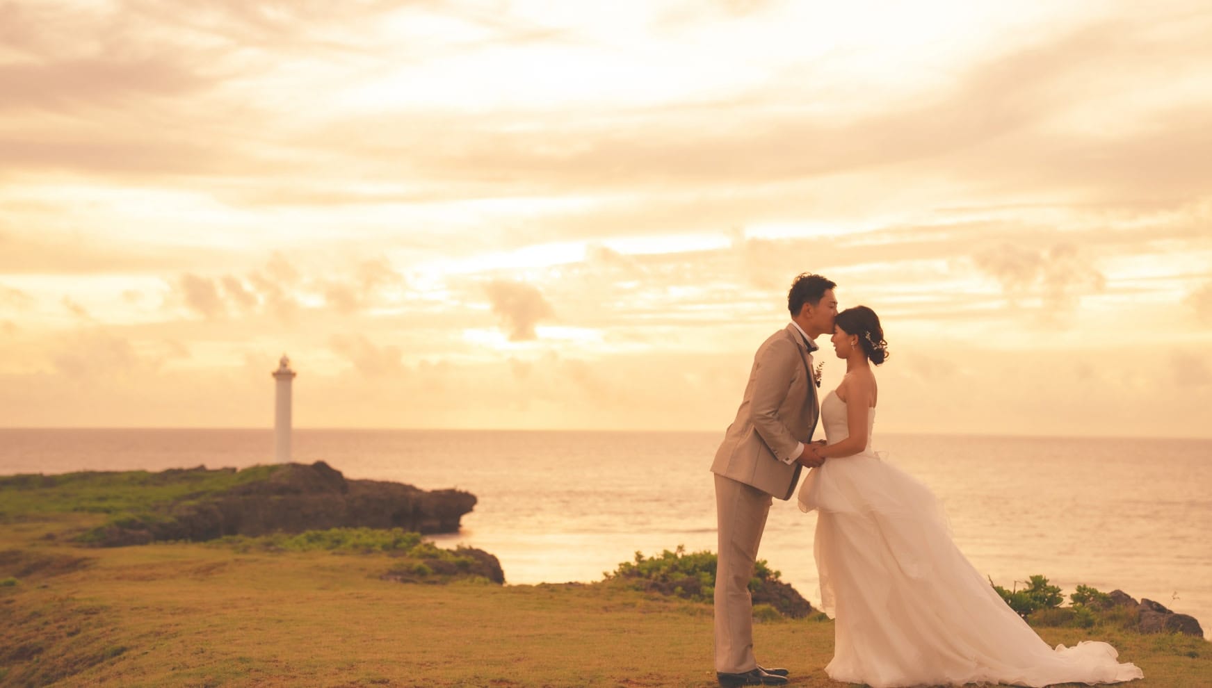沖縄本島の残波岬にて、黄金色に染まる夕日を背景に、新郎が新婦の額に優しくキスをするロマンチックなウエディングフォト。遠くに白い灯台を望む断崖の上で、逆光に照らされた二人のシルエットが美しい、感動的なサンセットロケーション撮影。