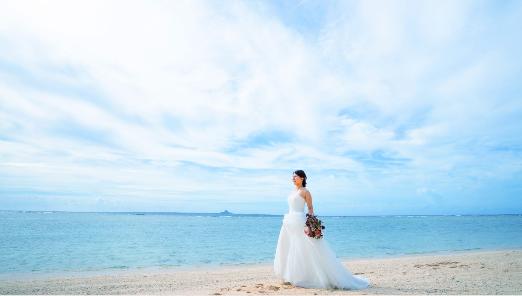 沖縄・本島北部の瀬底ビーチで撮影された、フォトウエディングのソロショット。透き通るようなエメラルドブルーの海と、どこまでも続く白い砂浜、広大な空を背景に、純白のウエディングドレスを纏った新婦が佇むドラマチックな一枚。
