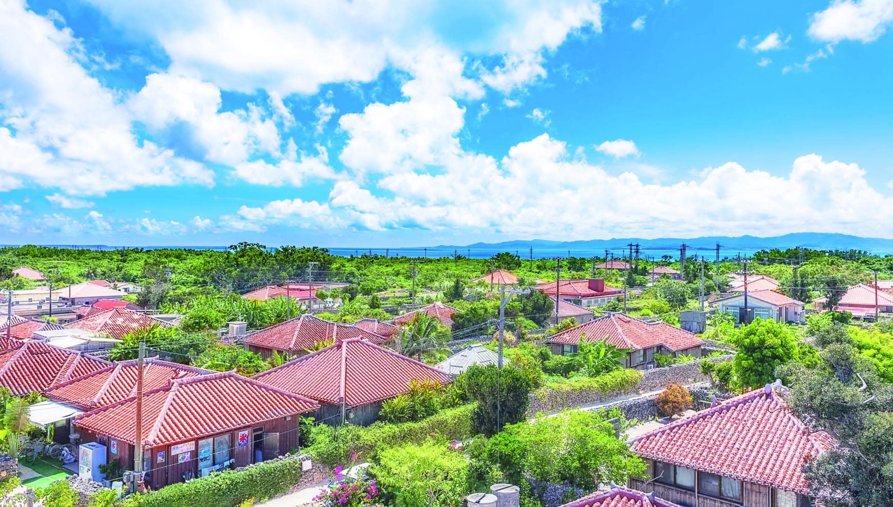 沖縄県・竹富島の伝統的な赤瓦屋根の集落を見渡す絶景。青い空と白い雲、周囲を囲む豊かな緑と美しい海が、重要伝統的建造物群保存地区ならではの情緒ある風景を彩る。沖縄の歴史と文化を感じるロケーションフォトの魅力を伝える一枚。