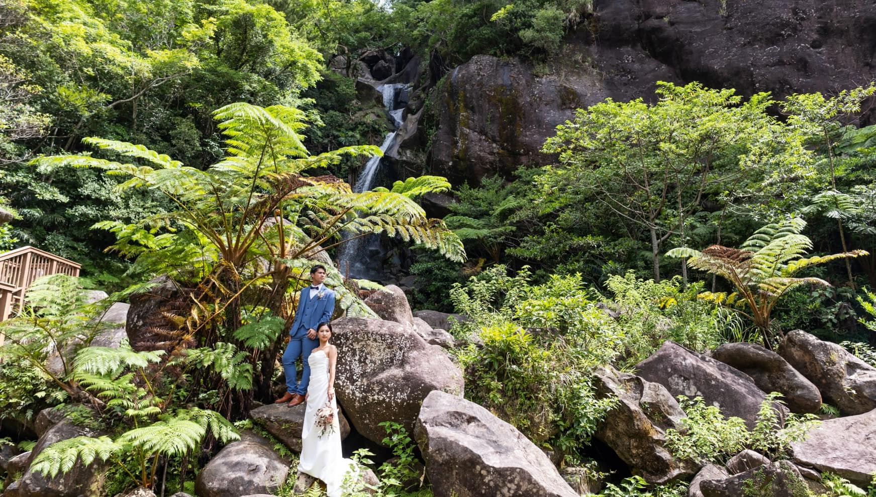 沖縄県名護市の「轟の滝（とどろきのたき）」を背景にしたドラマチックなフォトウエディング。力強く流れ落ちる滝と、太古の自然を感じさせる巨大なシダ植物に囲まれ、岩場に立つタキシード姿の新郎とウエディングドレス姿の新婦。