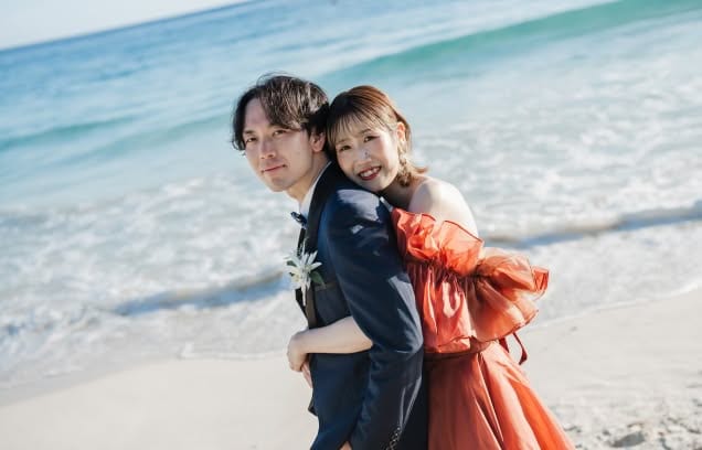 夕暮れ時のビーチで、紺色のタキシードを着た新郎の背中に、テラコッタオレンジのカラードレスを纏った新婦が優しく寄り添うバックハグのショット。波打ち際の穏やかな海を背景に、新婦の弾けるような笑顔が印象的な、ファッショナブルで温かみのあるウエディングフォト。