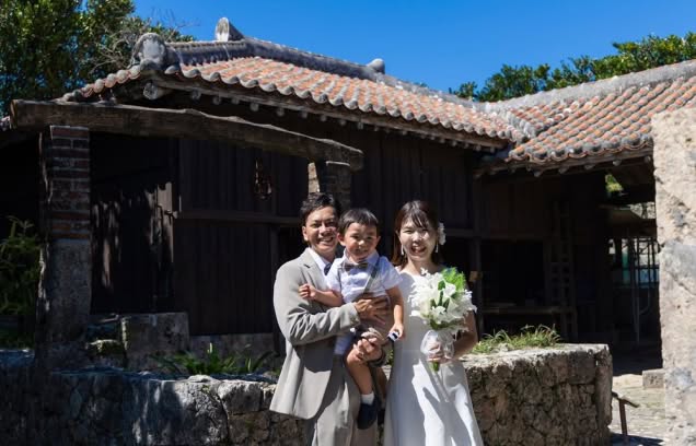 沖縄の伝統的な赤瓦屋根の古民家を背景に、お子様を抱っこして満面の笑みを見せる新郎新婦。歴史を感じさせる石垣と木造建築が、家族の温かみと沖縄らしい情緒を演出する、パパママ婚（ファミリーフォト）にぴったりのウエディングショット。
