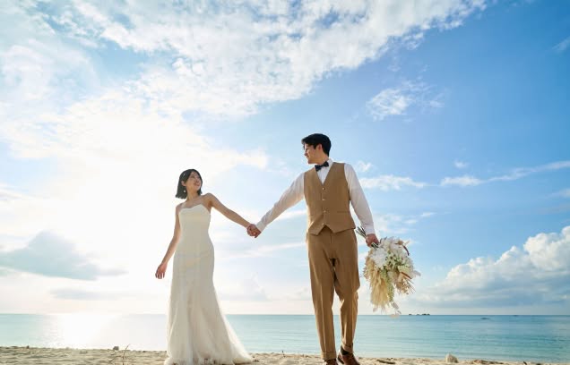 夕暮れ時の穏やかなビーチを、仲良く手を繋いで歩く新郎新婦。逆光気味の柔らかな光の中、キャメルカラーのベストとパンツを着こなした新郎と、マーメイドラインのドレスを纏った新婦が、お互いを見つめ合いながら進む、開放感と多幸感に満ちたウエディングフォト。
