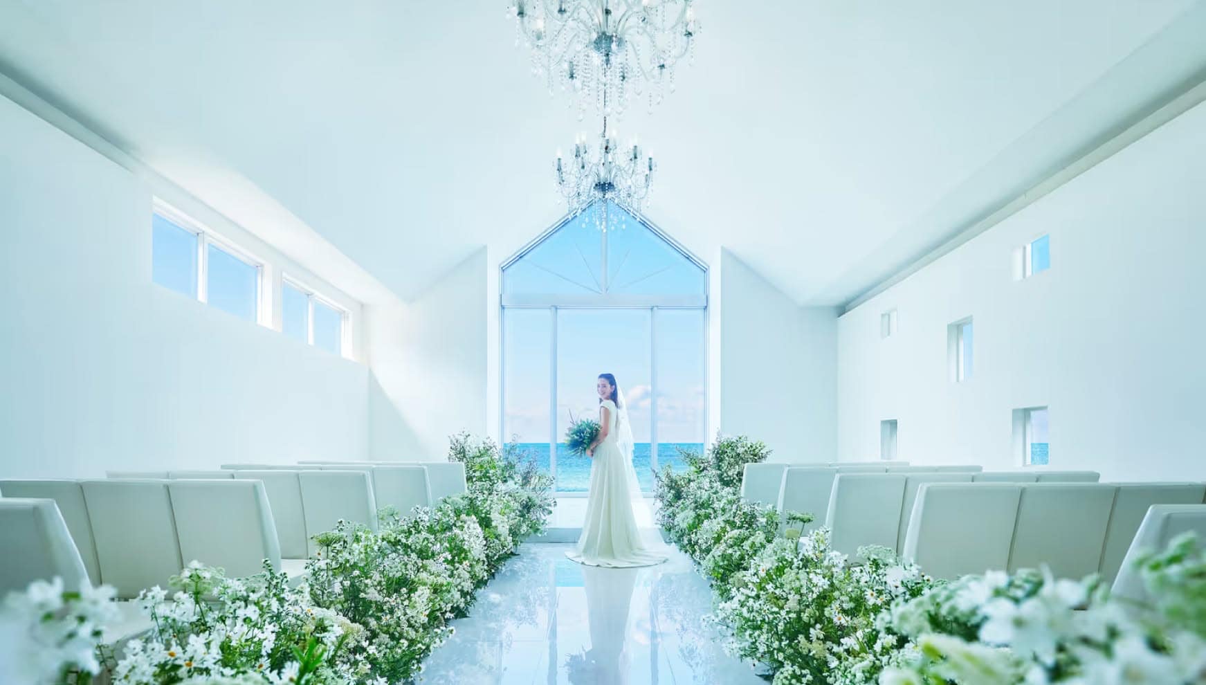 青い海が一望できる全面ガラス張りの白亜のチャペルに、一人佇むウエディングドレス姿の花嫁。高い透明感と自然光が美しい空間で、バージンロードの装花がリゾートウエディングの清らかな雰囲気を引き立てています。