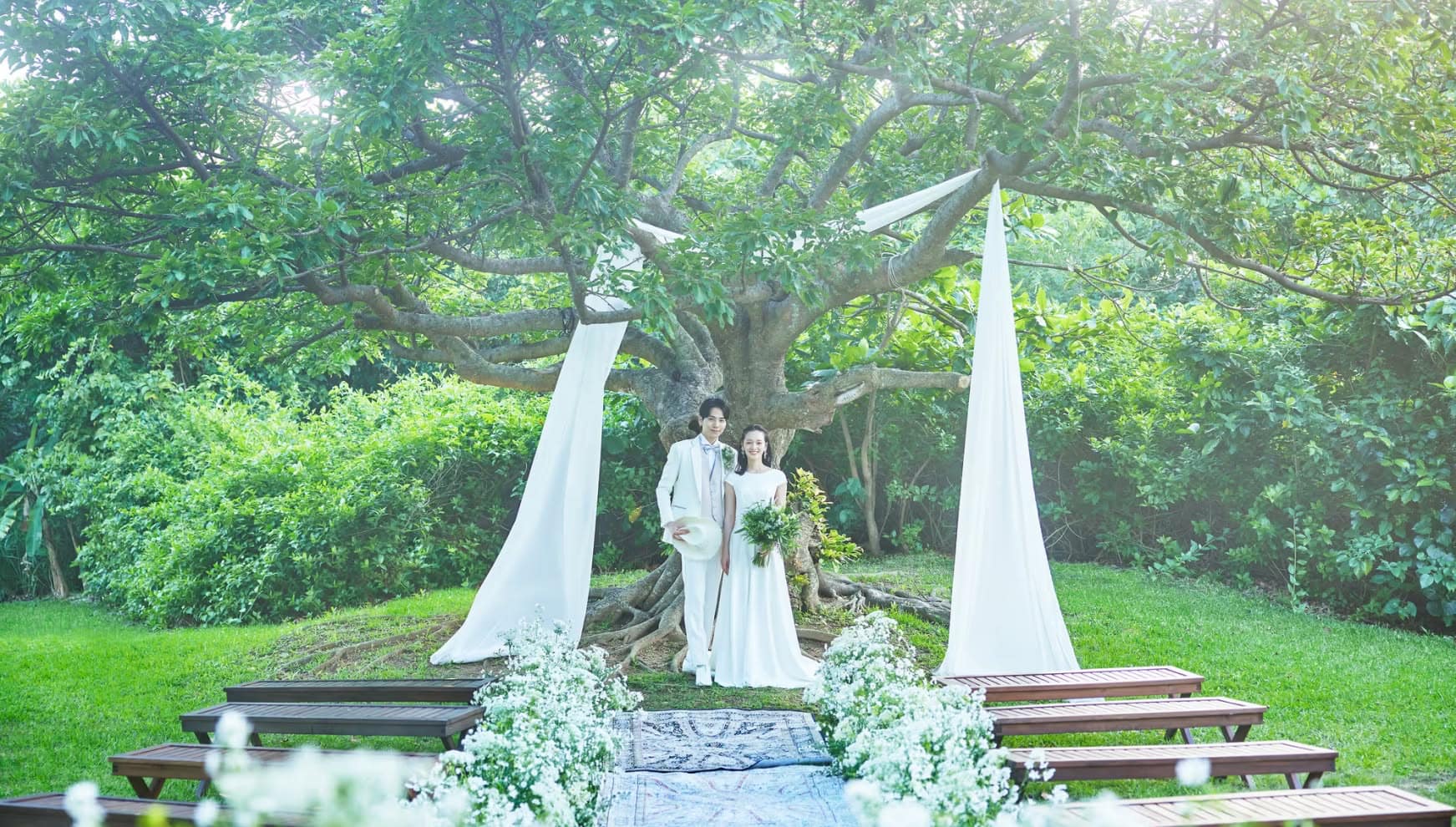石垣島の豊かな緑に囲まれたガーデンにて、大きなガジュマルの木を背に立つ新郎新婦。白いウエディングドレスとタキシード姿の二人が、木から吊るされた白い装飾布と白い花々に彩られたバージンロードの先で、穏やかに微笑むナチュラルなウエディングシーン。