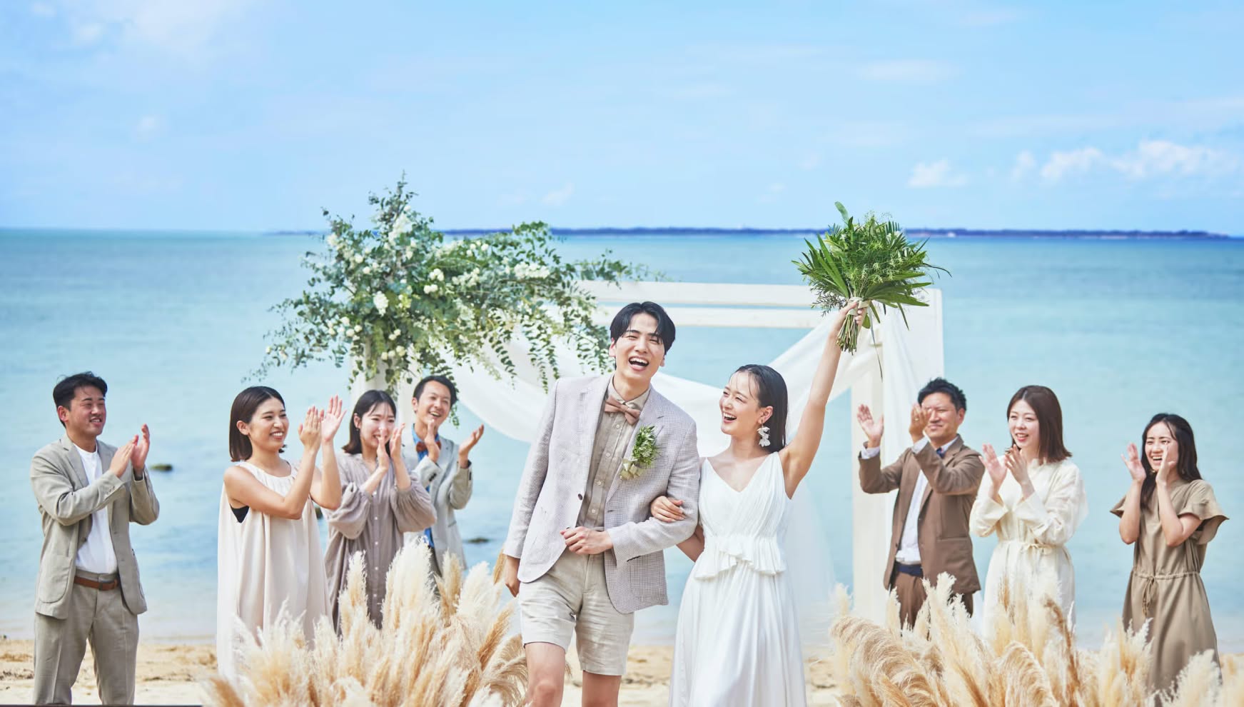 石垣島の透き通るような青い海を背景に、ビーチウエディングの退場シーンで笑顔を見せる新郎新婦。ゲストからの温かい拍手に包まれながら、花嫁がブーケを高く掲げ、リゾートらしい開放感あふれる雰囲気の中で祝福を受ける幸せな瞬間の様子。