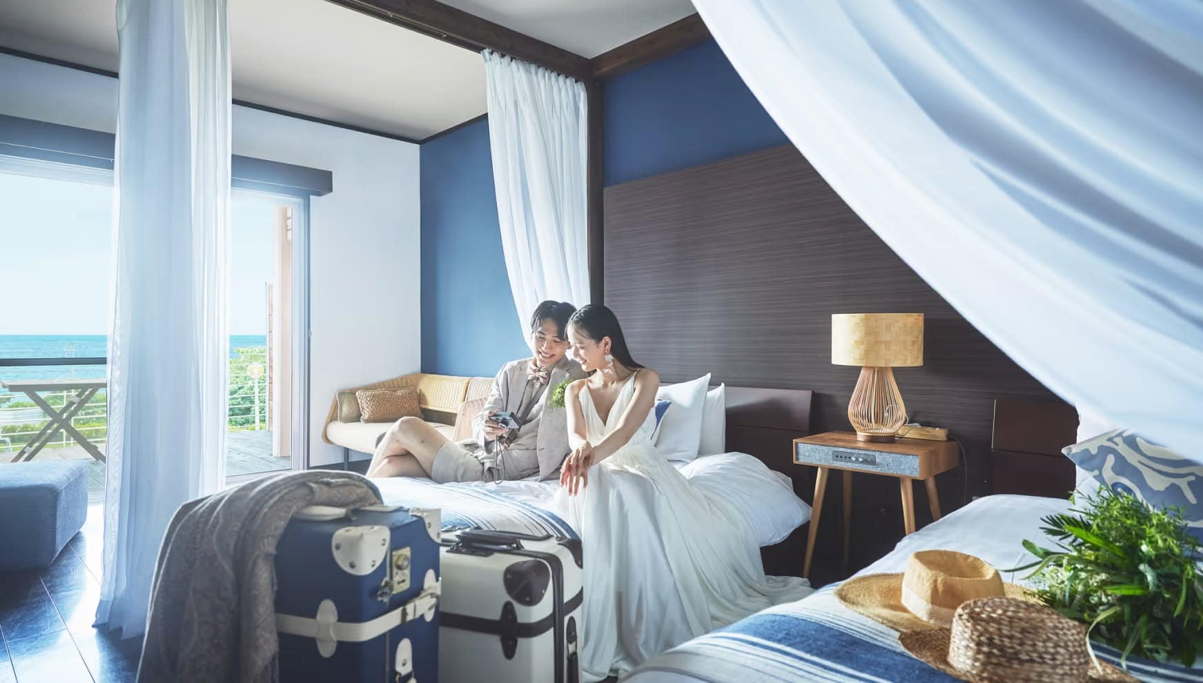 石垣島の海が見えるホテルの客室にて、ウエディングドレスとタキシード姿のままベッドに座り、カメラを眺めて微笑む新郎新婦。ブルーの壁がアクセントのモダンな部屋には、トランクや麦わら帽子が置かれ、挙式後のプライベートな時間を楽しむリラックスした雰囲気。