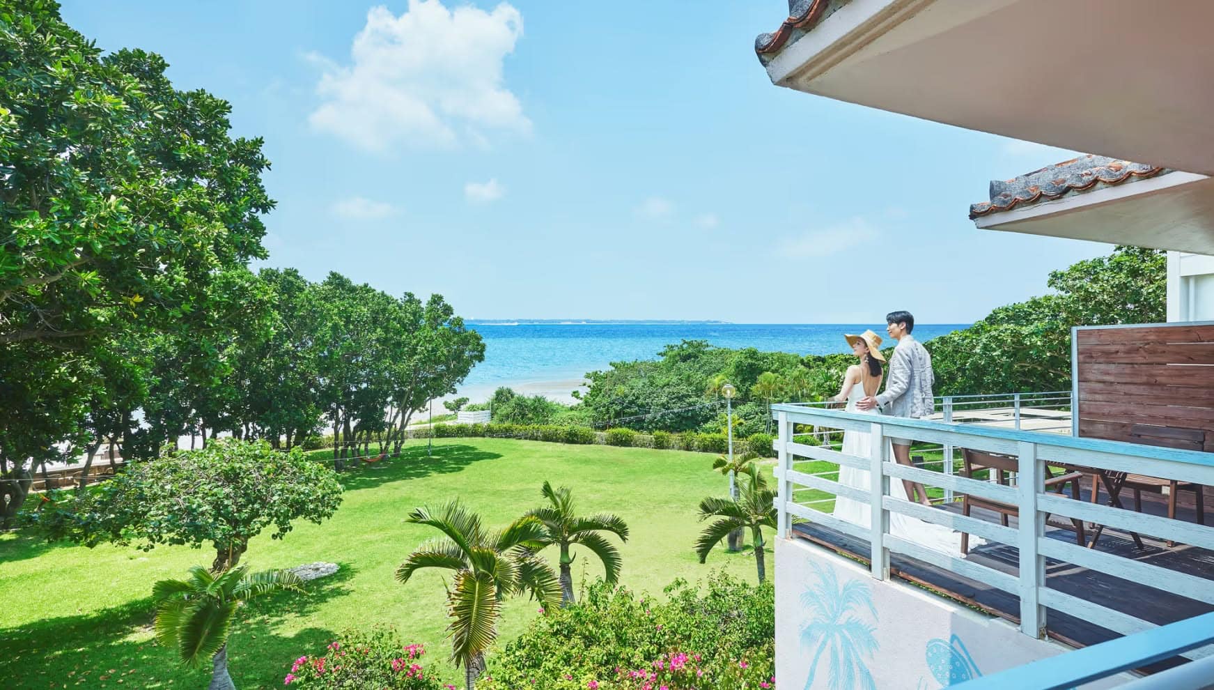 石垣島のザ・ビーチテラス ホテル・アオのテラスから、エメラルドグリーンの海を眺めるウエディング姿のカップル。麦わら帽子を被った新婦とグレーのジャケットを着た新郎が、南国の木々や芝生が広がるガーデンを見下ろす、開放感あふれるリゾートの風景。