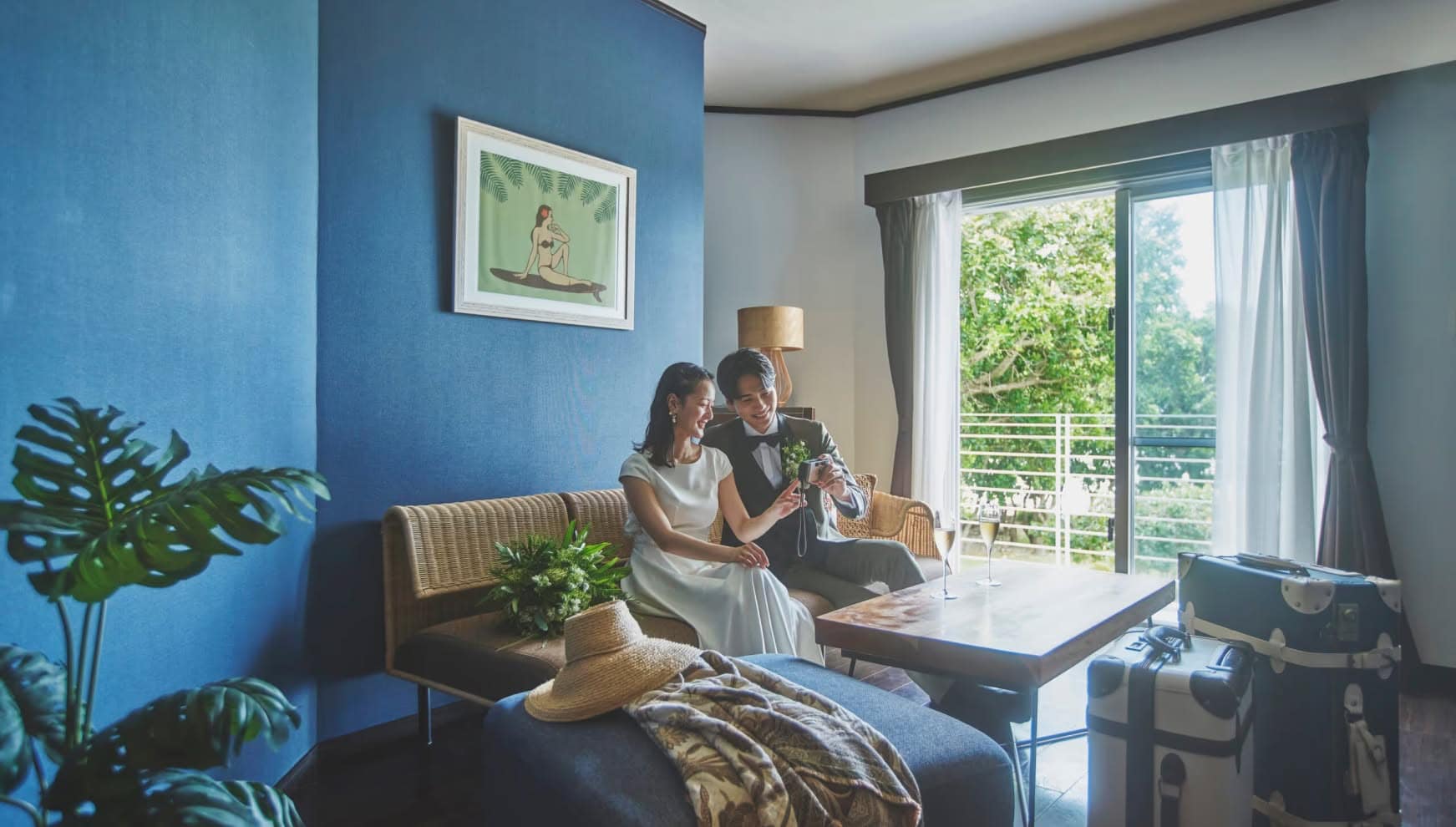 石垣島のホテルの客室にて、青いアクセントウォールの前にあるラタンソファに座り、二人でデジタルカメラの画面を覗き込む新郎新婦。テーブルにはシャンパングラスが置かれ、室内にはモンステラの観葉植物やトランクが並ぶ、挙式後の穏やかなひととき。
