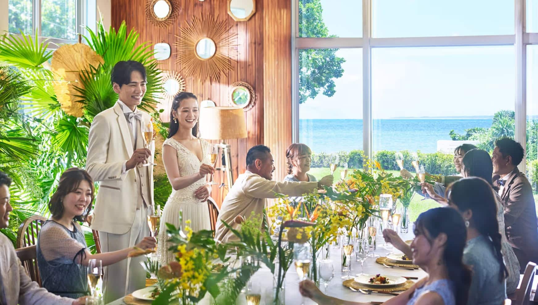 石垣島の海を一望できるオーシャンビューのパーティー会場にて、新郎新婦とゲストがシャンパングラスを掲げて乾杯する様子。木目調の壁や鮮やかなグリーンの装飾に囲まれた空間で、ウエディングドレスとタキシード姿の二人が笑顔でゲストと交流する祝宴の風景。