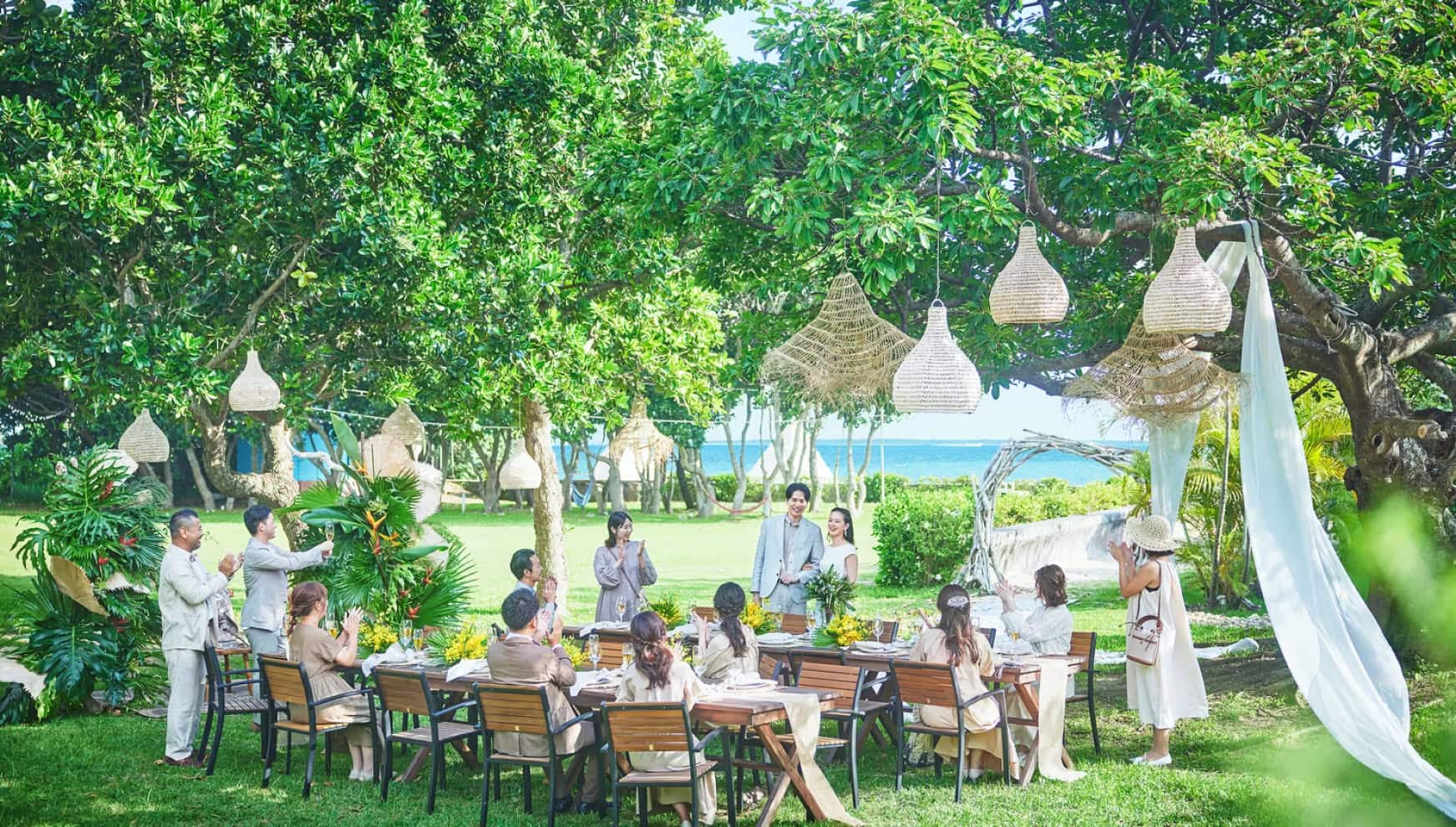 石垣島の自然に囲まれたガーデンにて、大きな木に吊るされたラタンのランプシェードが印象的な屋外パーティー会場。新郎新婦を囲み、長テーブルに座るゲストが拍手で祝福する様子。背景には青い海が広がり、リゾートならではの開放感あふれるウエディングパーティーの風景。