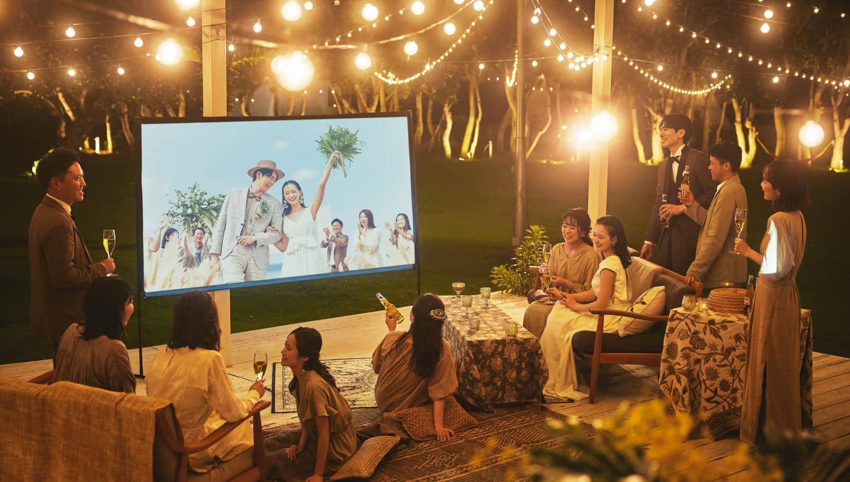 夜の石垣島のガーデンにて、ライトアップされた幻想的な雰囲気の中で行われるウエディングパーティー。新郎新婦とゲストが屋外のスクリーンで思い出の映像を鑑賞しながら、お酒を片手に談笑する、リゾートならではのナイトパーティーの風景。