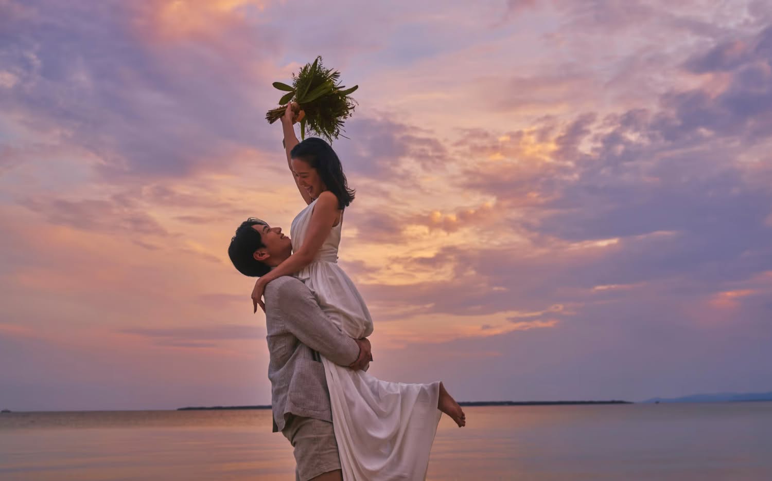 沖縄の海辺で、夕焼け空を背景に新郎が新婦を抱きかかえるドラマチックなサンセットフォト。新婦はブーケを高く掲げ、二人のシルエットがオレンジ色の空に映える、幻想的で幸福感あふれるウエディングのロケーション撮影です。