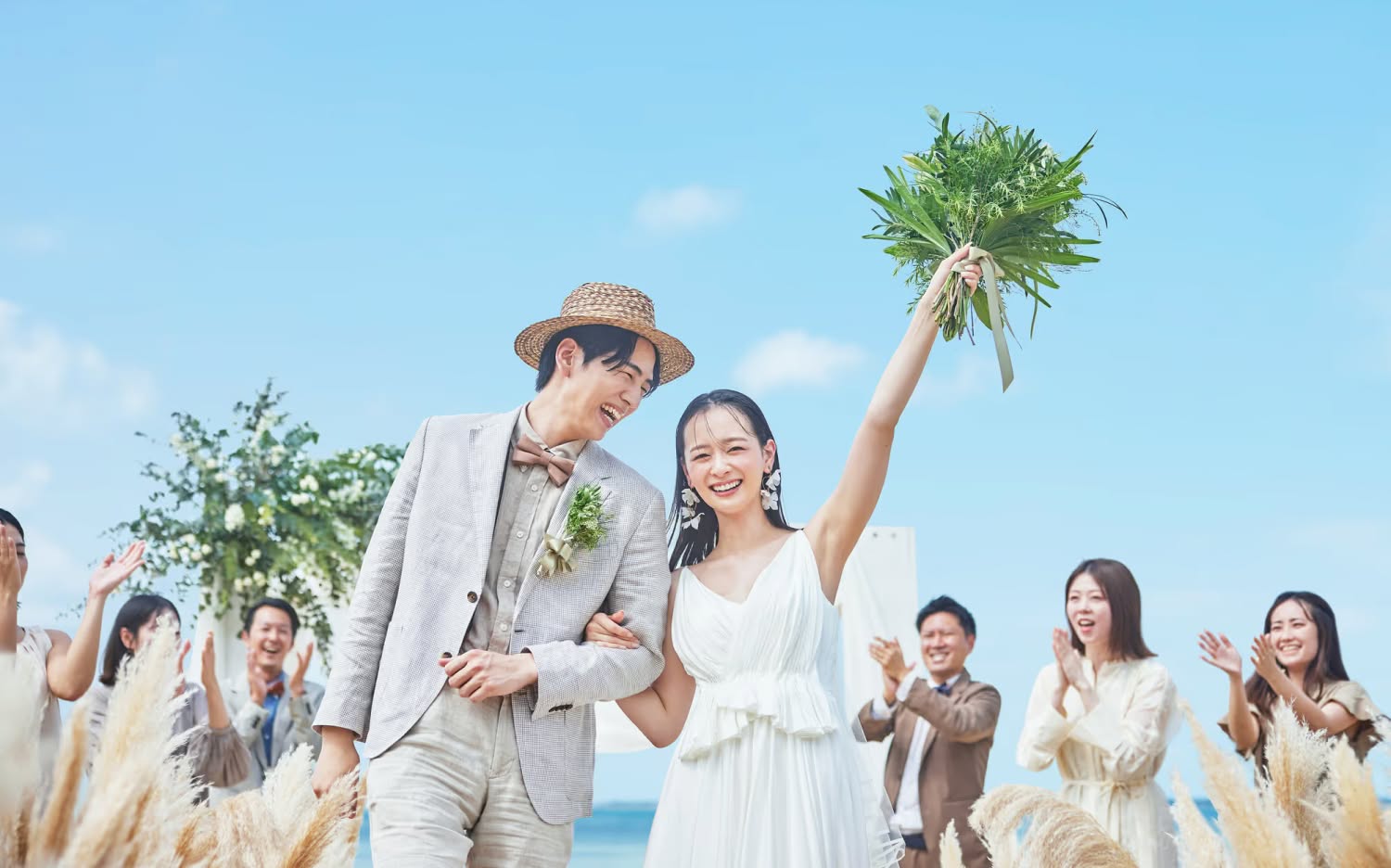青い海を背景にしたビーチウエディングで、幸せそうな新郎新婦と、二人を笑顔で見守る参列者たち