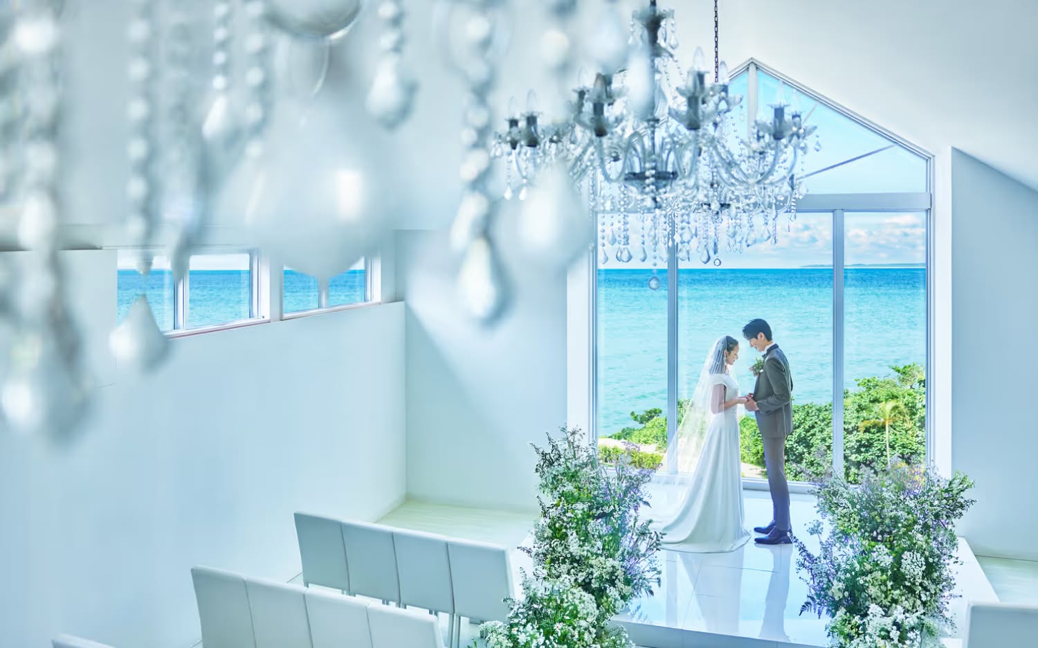 沖縄の海を一望できる白亜のチャペルで、手を取り合うウエディング姿の新郎新婦。クリスタルのシャンデリアが輝く開放的な空間と、窓の外に広がるエメラルドグリーンの海が幻想的な、リゾートでの挙式を象徴する一枚です。