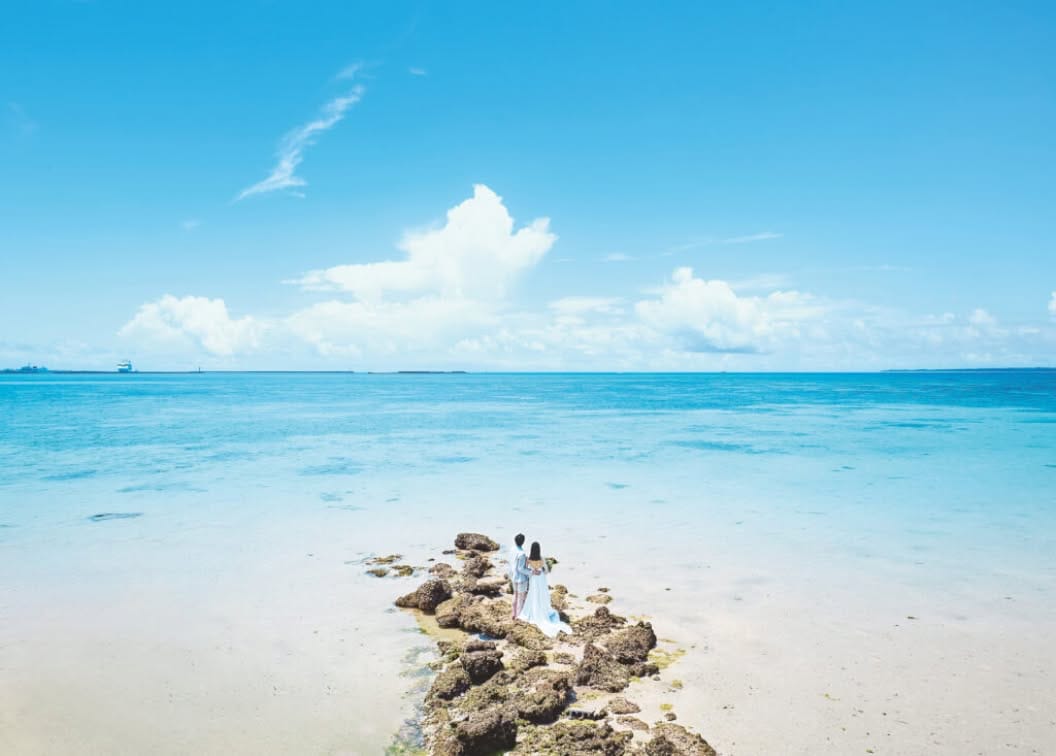 石垣島の透き通るようなエメラルドグリーンの海を背景に、浜辺の岩場に寄り添って立つ新郎新婦のバックスタイル。どこまでも続く青い空と白い雲が広がる開放的なロケーションで、ウエディングドレスの裾をなびかせながら絶景を眺めるドラマチックな風景。