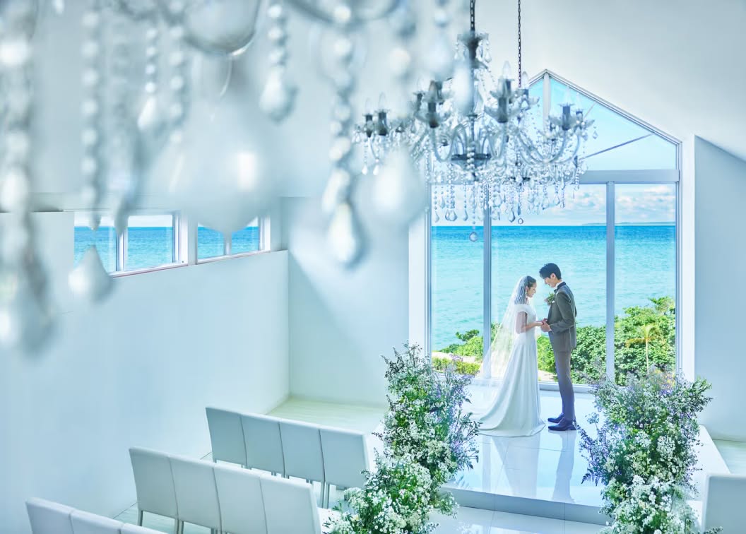 沖縄の海を一望できる白亜のチャペルで、手を取り合うウエディング姿の新郎新婦。クリスタルのシャンデリアが輝く開放的な空間と、窓の外に広がるエメラルドグリーンの海が幻想的な、リゾートでの挙式を象徴する一枚です。