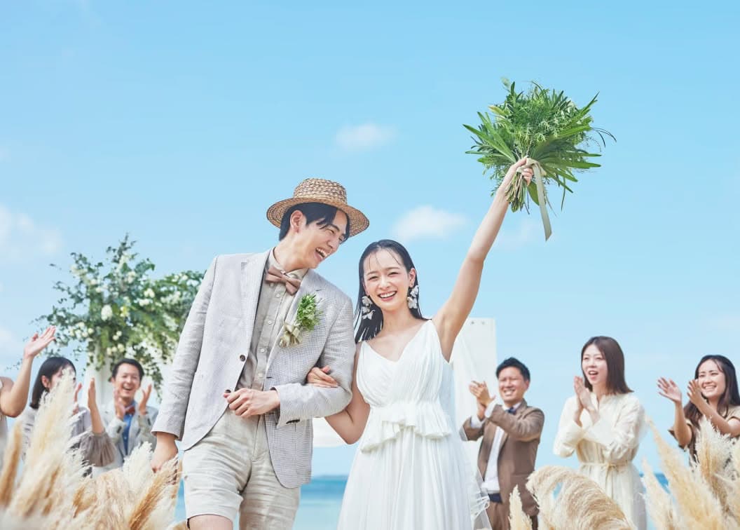 青い海を背景にしたビーチウエディングで、幸せそうな新郎新婦と、二人を笑顔で見守る参列者たち