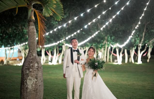 夜の石垣島のガーデンにて、頭上に張り巡らされたイルミネーションが美しく輝く中、腕を組んで微笑む新郎新婦。白いタキシードと刺繍が施されたウエディングドレス姿の二人が、南国の木々に囲まれた幻想的な空間で佇む、ナイトウエディングの華やかな風景。
